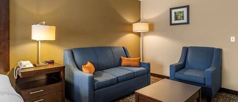 A hotel room with a blue sofa, a matching armchair, a wooden nightstand with a phone, and a lamp.