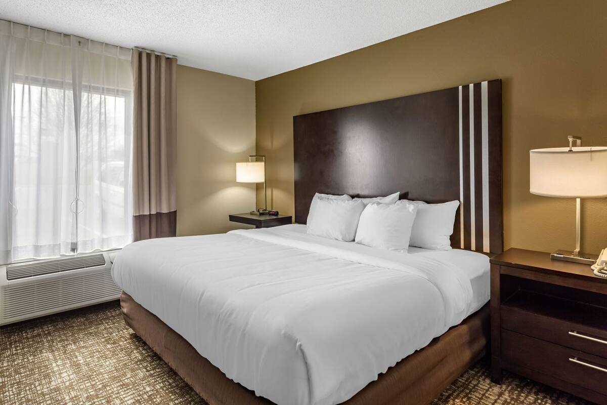 A hotel room with a large bed, a nightstand with a lamp, a wall-mounted telephone, and a window with curtains.