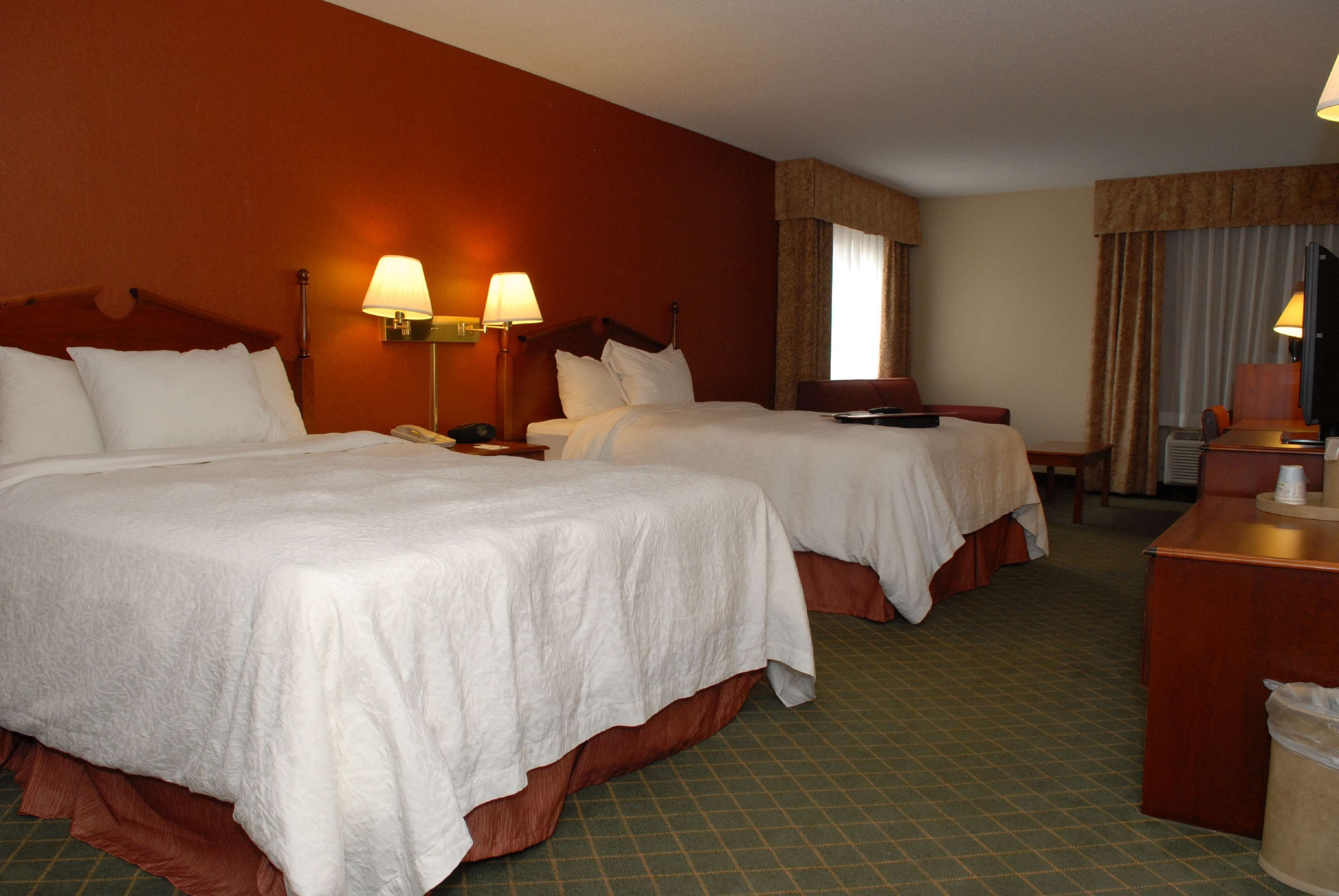 Une chambre d’hôtel avec deux lits, un bureau et une chaise. Il y a des lampes au mur et une fenêtre avec des rideaux.