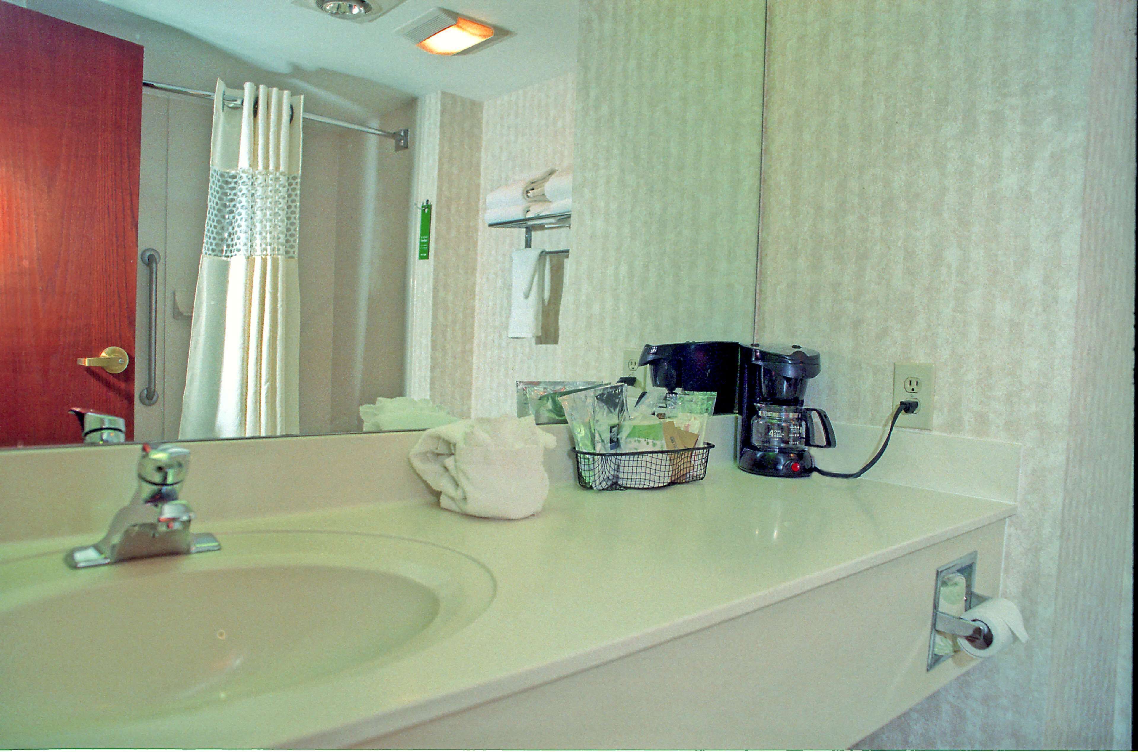 A bathroom with a sink, a mirror, a towel, a basket of toiletries, and a coffee maker.
