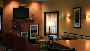 Une chambre d’hôtel avec une table à manger en bois, des chaises rouges et beiges, un téléviseur fixé au mur, une fenêtre avec des rideaux et des lampes murales.