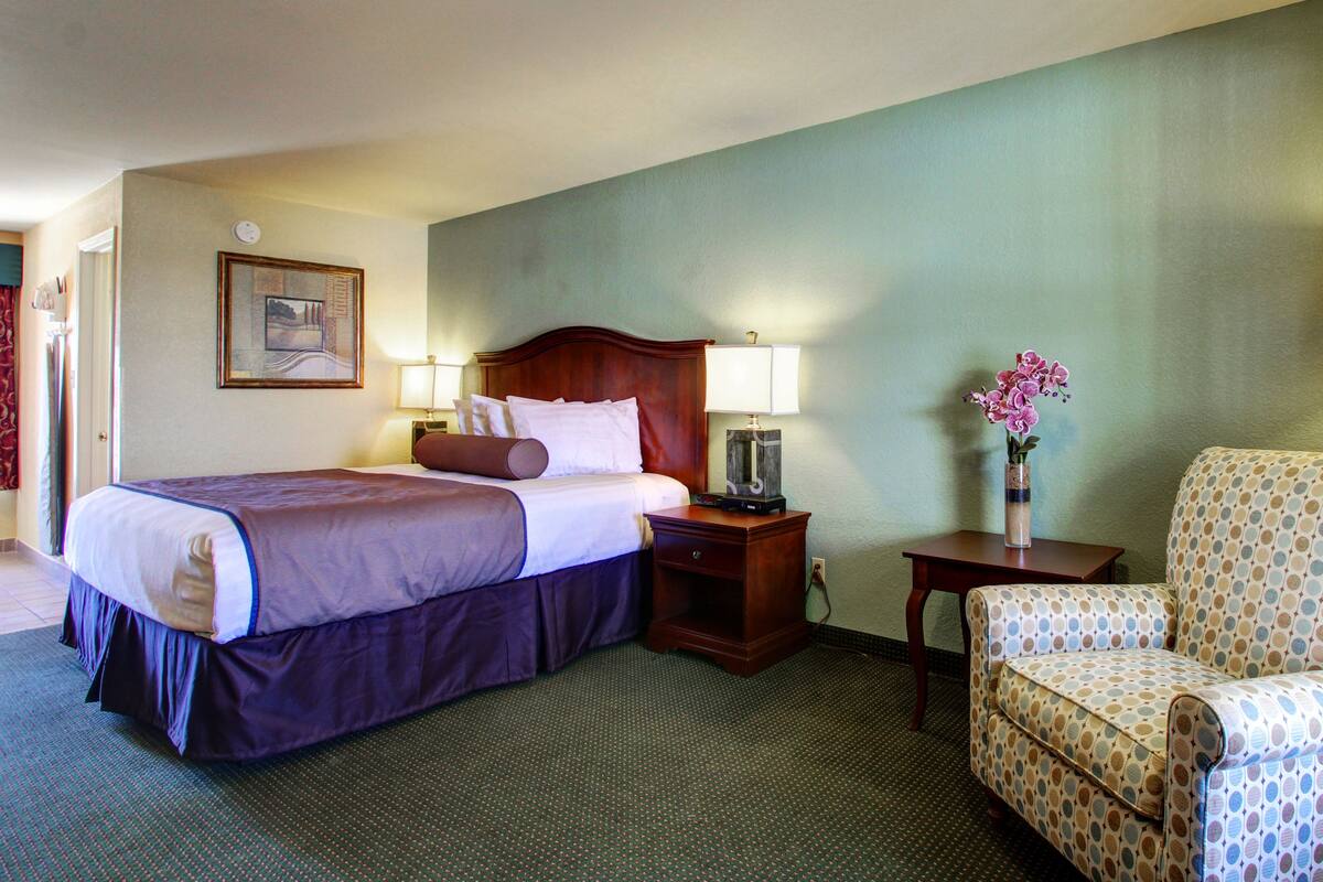 A hotel room with a bed, a nightstand, a chair, and a wall clock.