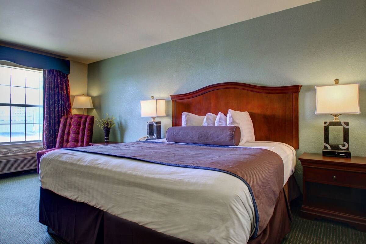 A hotel room with a large bed, two bedside lamps, a chair, a window with curtains, and a nightstand.