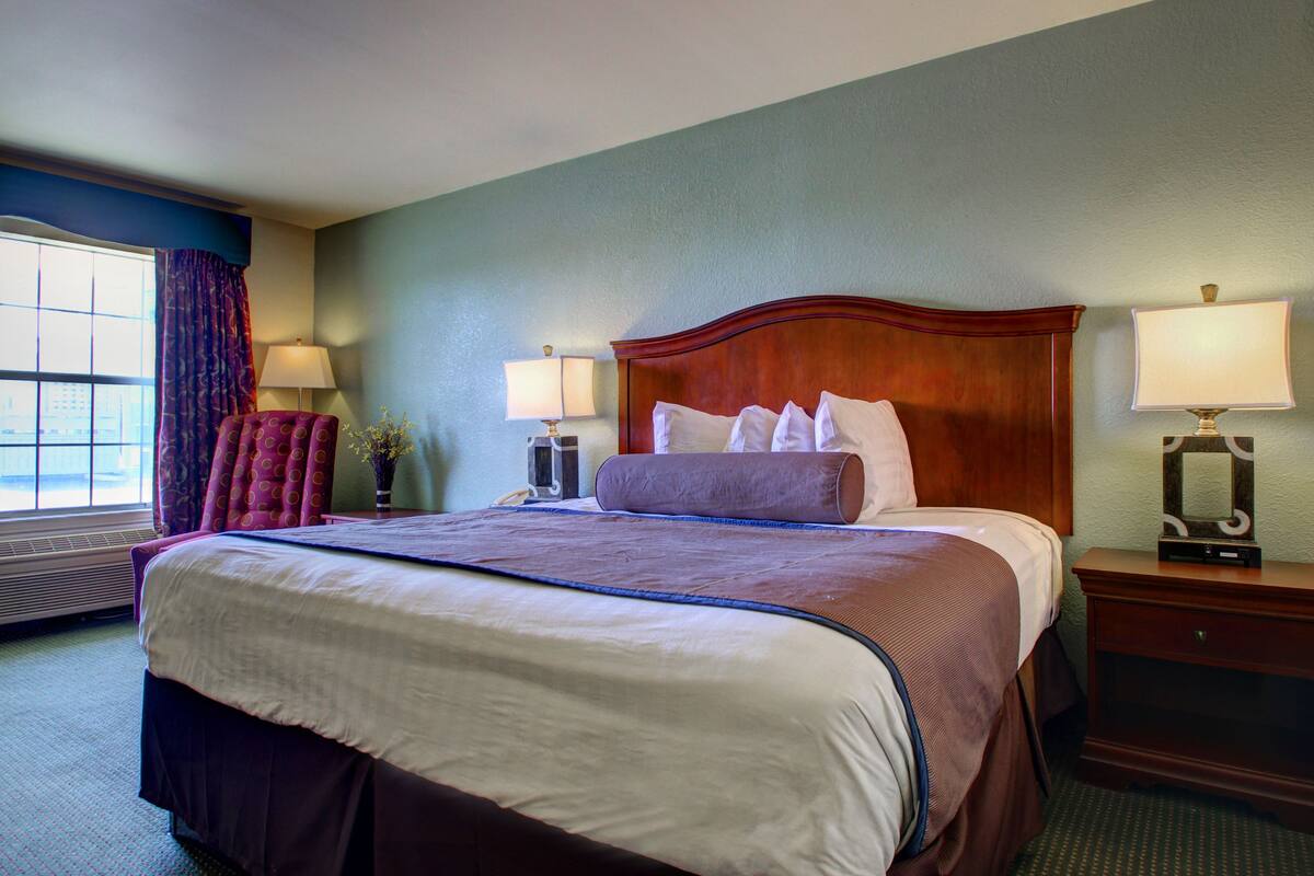 A hotel room with a large bed, two bedside tables with lamps, a chair, and a window with curtains.
