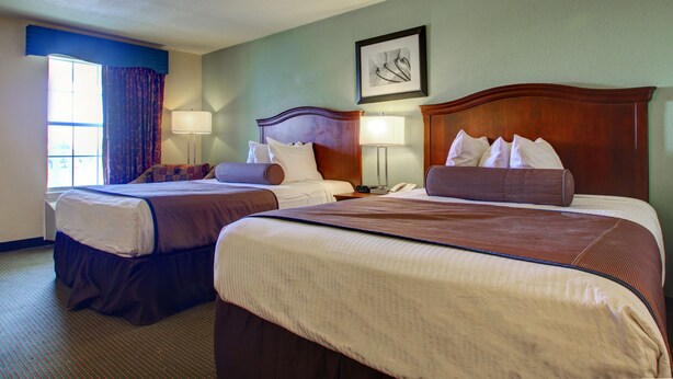 A hotel room with two beds, a large window with curtains, a framed artwork on the wall, and a lamp on each nightstand. - Key West Inn Tunica (Robinsonville)