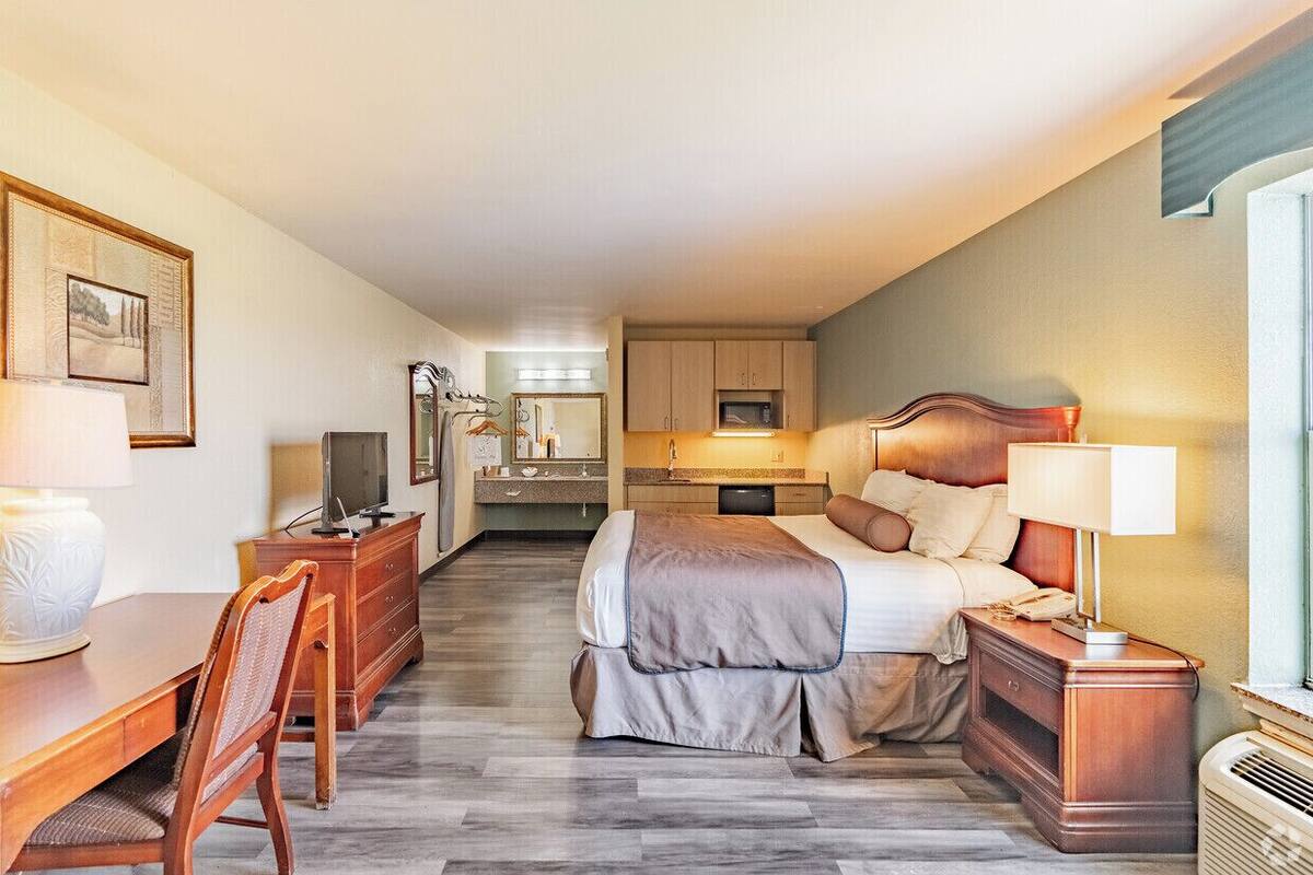 A hotel room with a bed, bedside table, desk, chair, and a kitchenette area.