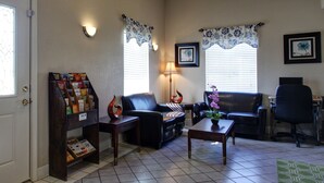 A living room with a black leather sofa, a coffee table, a bookshelf, and two windows with floral curtains. - Key West Inn Tunica (Robinsonville)