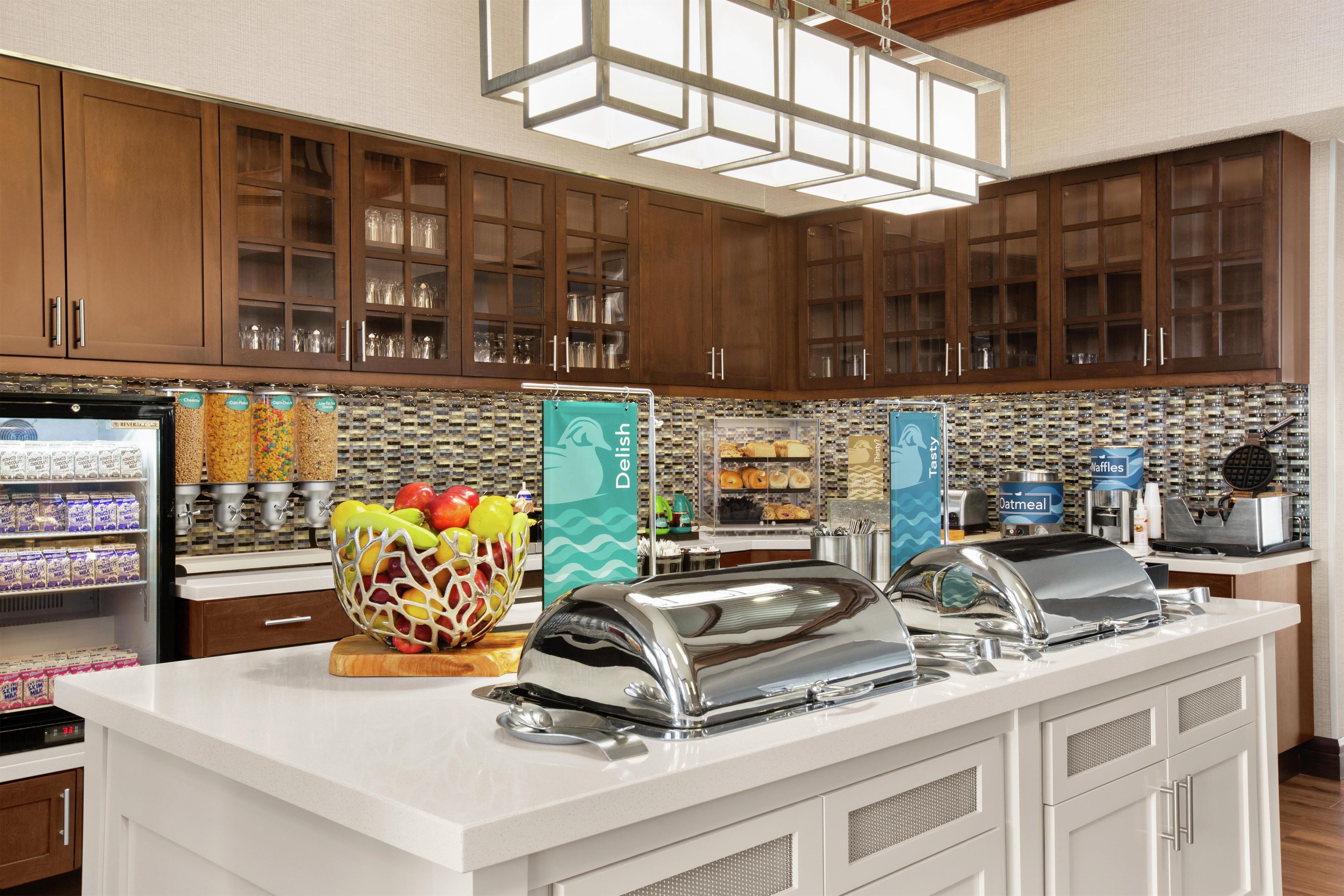 A hotel kitchen with a buffet setup, including a refrigerator with beverages, a fruit bowl, and a buffet counter with various food items.