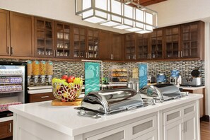 Una cocina de hotel con montaje tipo buffet, que incluye un refrigerador con bebidas, un cuenco de fruta y un mostrador buffet con diversas opciones de comida.
