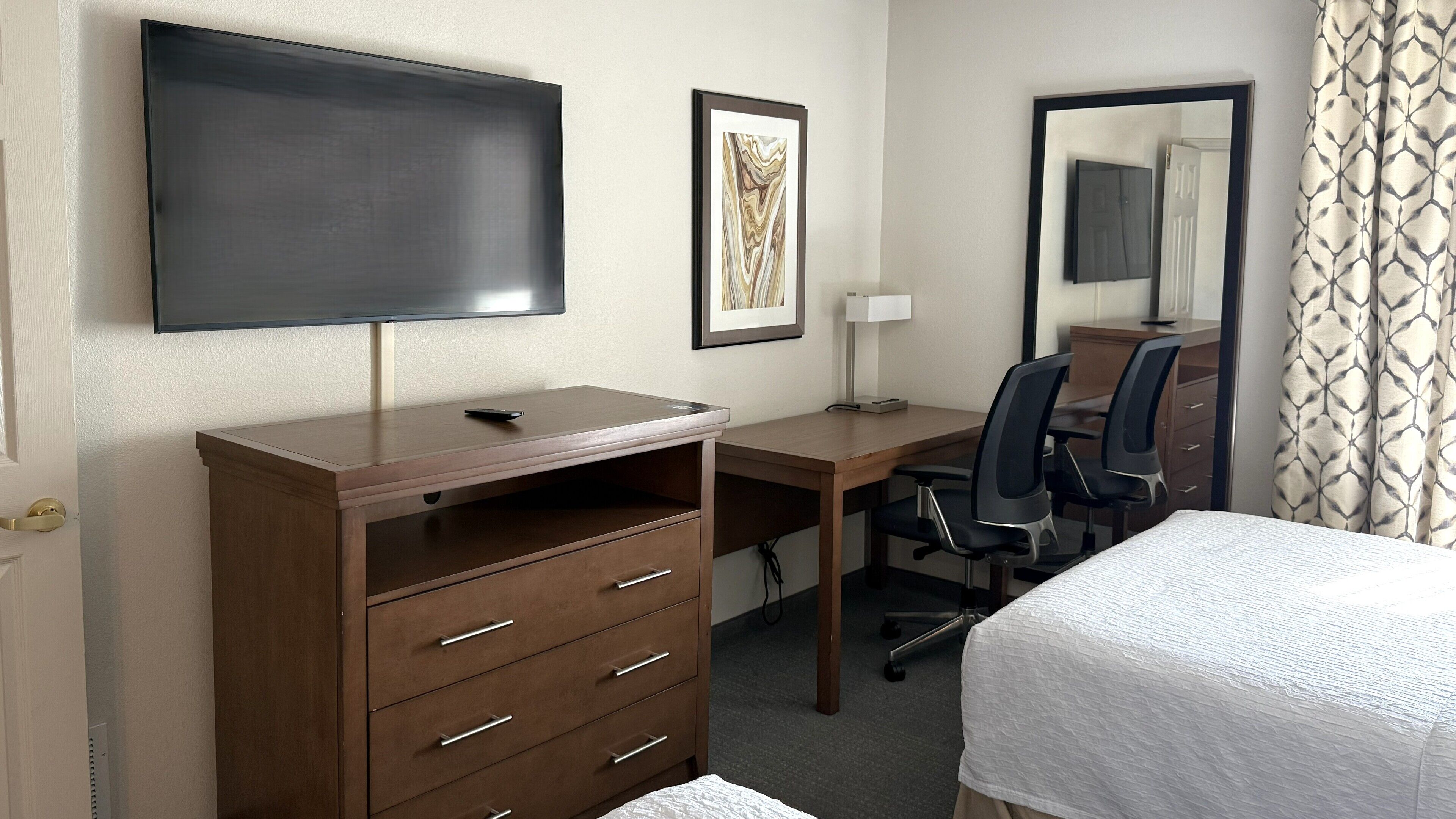 A hotel room with a flat-screen TV mounted on the wall, a desk with a chair, a bed with a white bedspread, and a framed artwork on the wall.