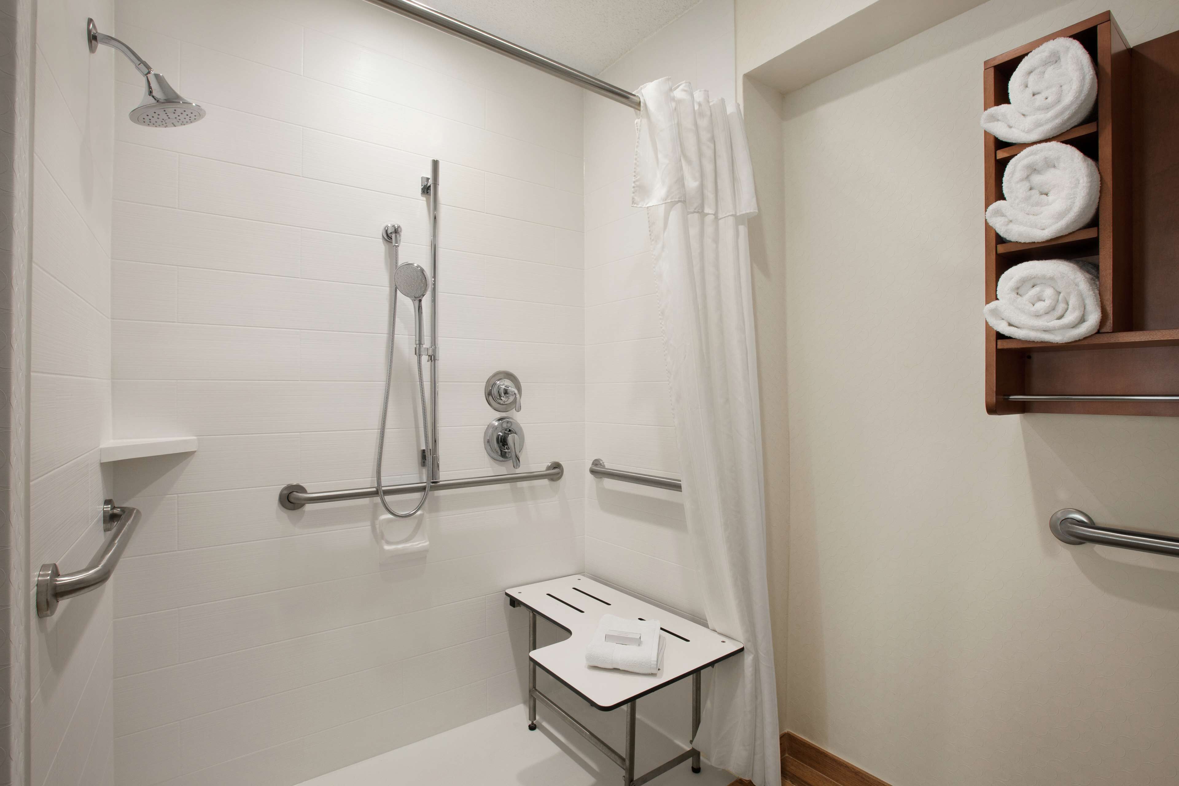 A modern bathroom with white tiles, a shower with a handheld showerhead, a glass-enclosed toilet, and a wooden shelf with rolled towels.