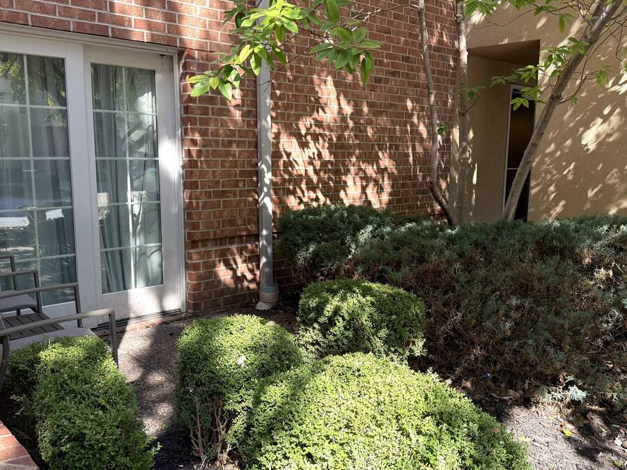 A brick building with a sliding glass door, a small patio with a table and chairs, and well-maintained bushes.