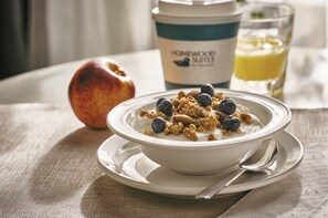 Un tazón de yogur con granola y arándanos sobre un plato blanco, una taza de café y un vaso de jugo de naranja en una mesa.