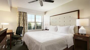 A hotel room with a bed, two bedside lamps, a desk with a chair, a ceiling fan, and a window with curtains.