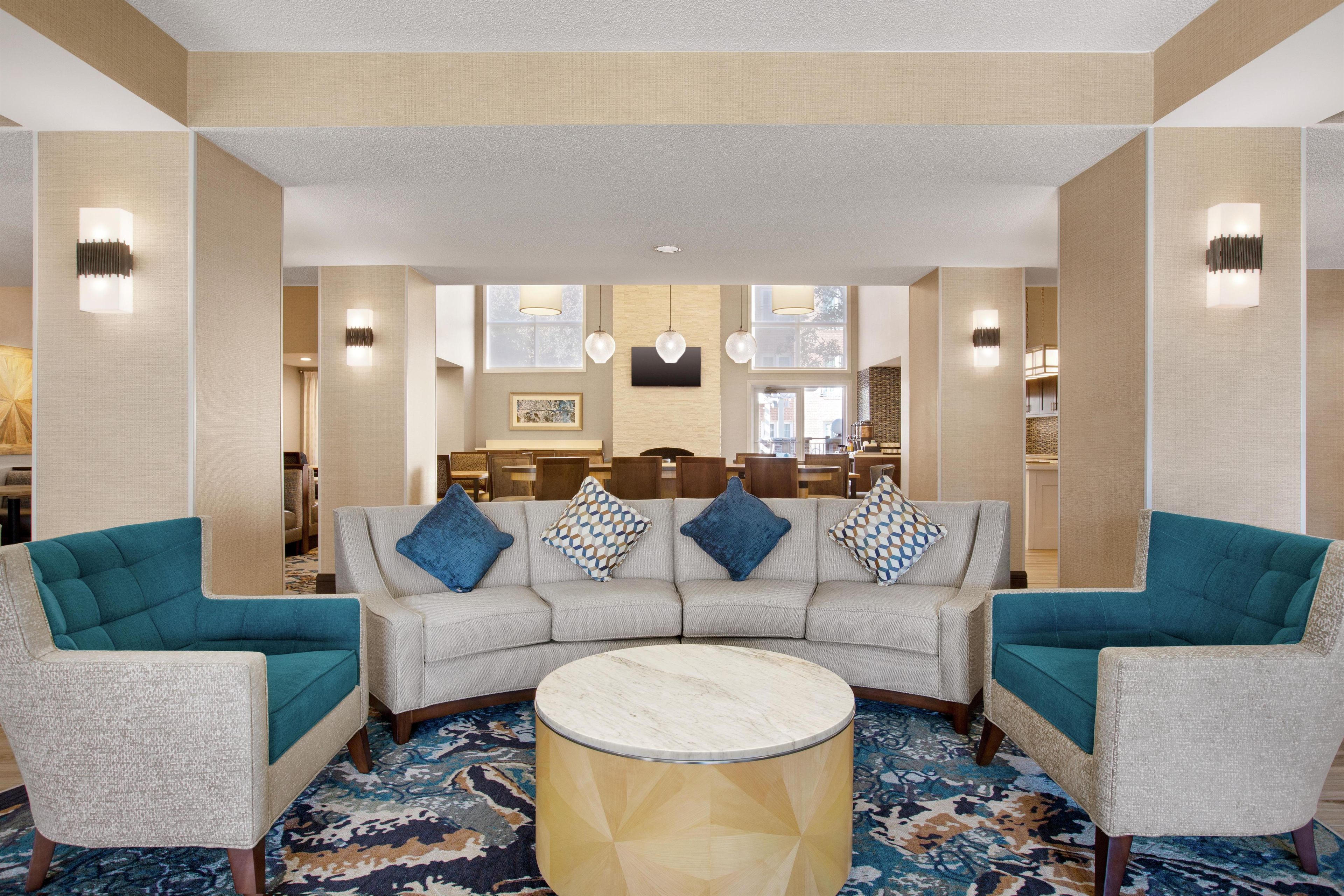 A hotel lobby with a curved grey sofa, teal armchairs, a round marble coffee table, and a patterned carpet.