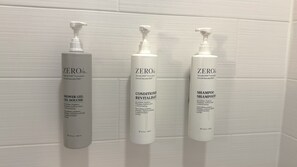 Tres botellas de champú, acondicionador y gel de ducha Zero% sobre una pared de azulejos blancos.