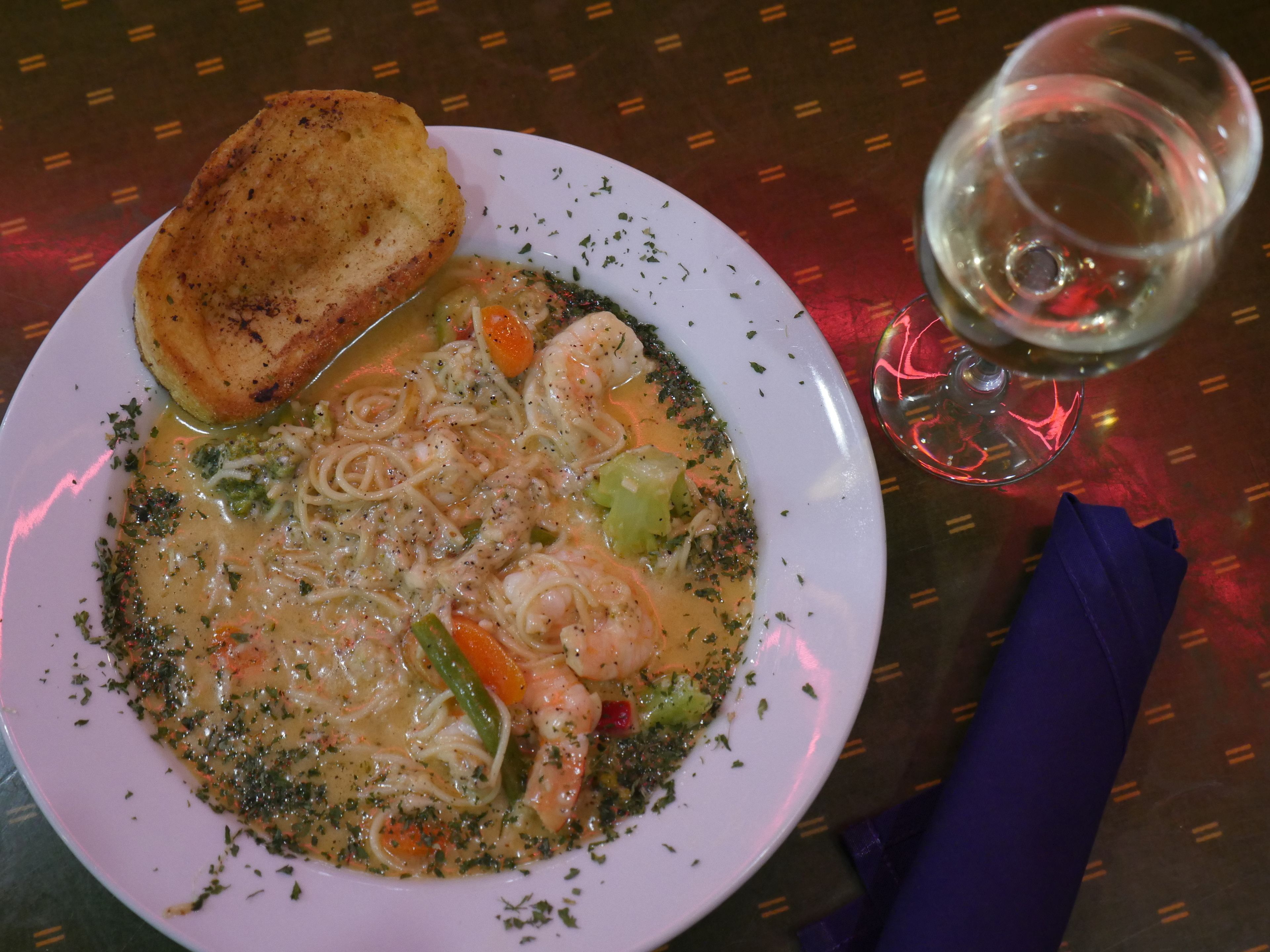 A plate of seafood pasta with vegetables and a toasted bread roll, accompanied by a glass of white wine on a table with a patterned tablecloth.