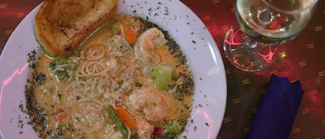 A plate of seafood pasta with vegetables and a toasted bread roll, accompanied by a glass of white wine on a table with a patterned tablecloth.