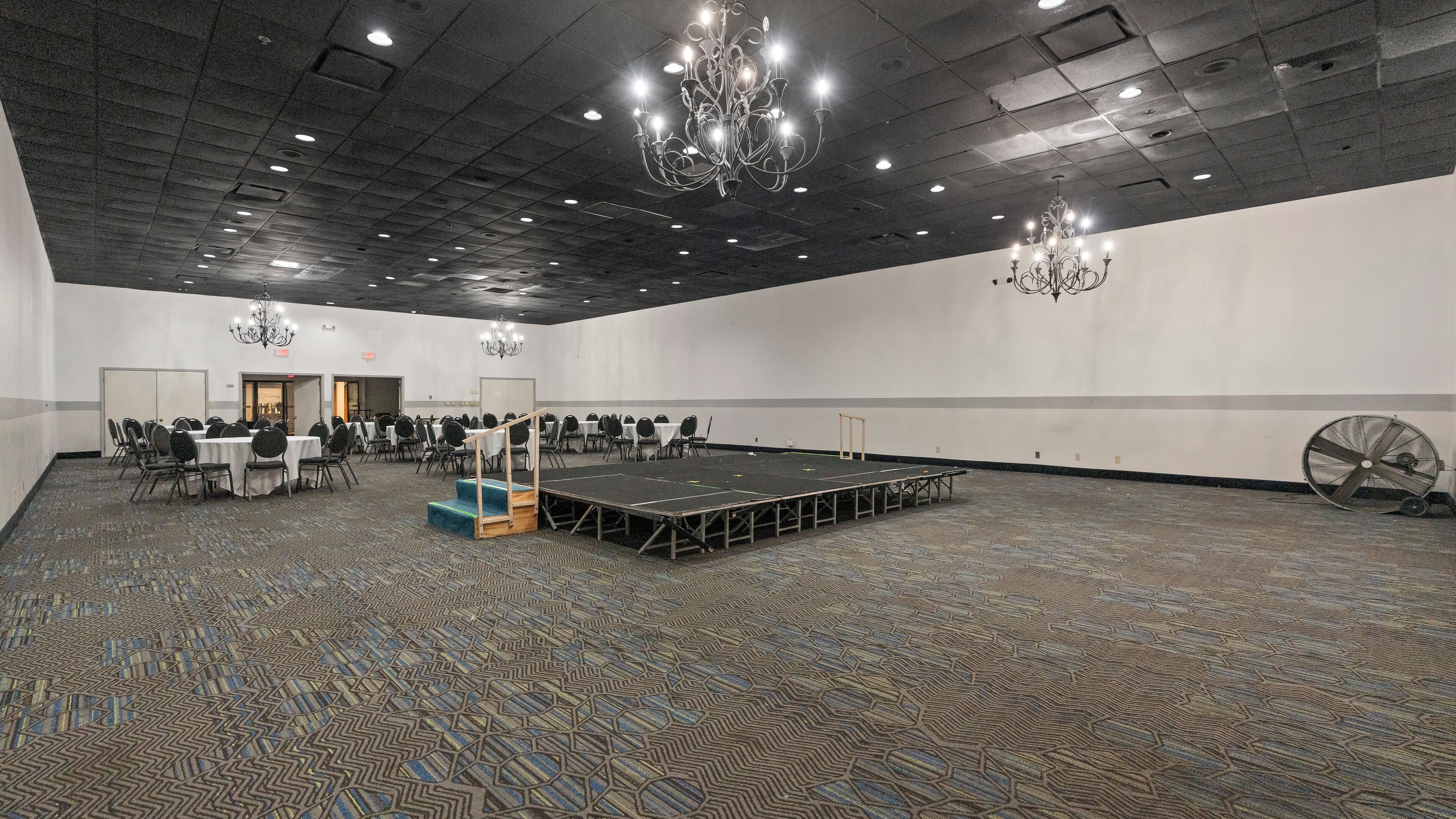 A large conference room with a stage, multiple tables, and chairs, equipped with chandeliers and a ceiling fan.