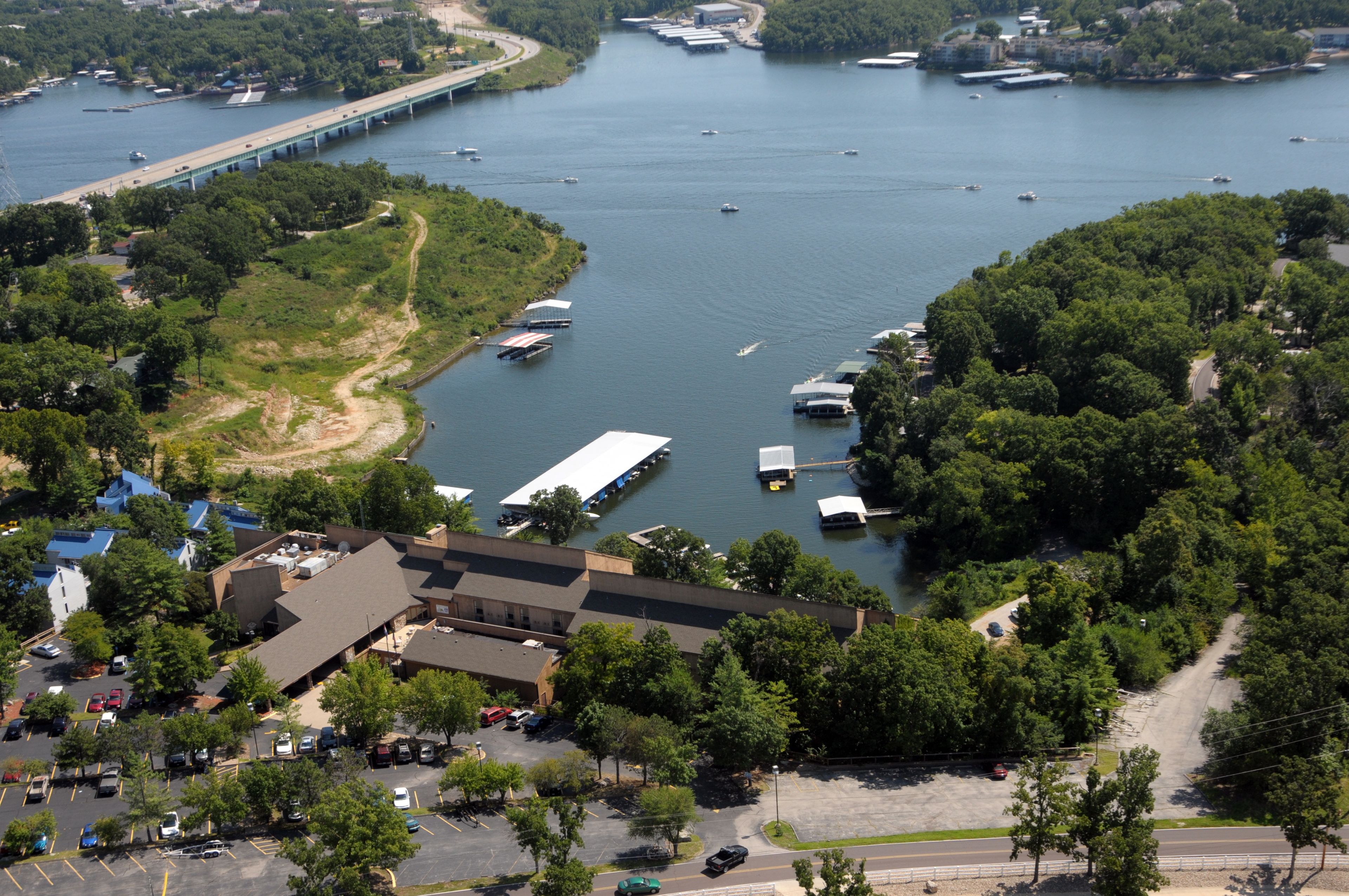 An aerial view of a waterfront property with a parking lot, a building with a sloped roof, and a bridge in the background.