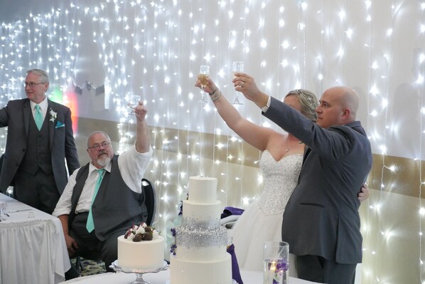 A wedding reception with a tiered white cake, guests toasting, and a backdrop of string lights.