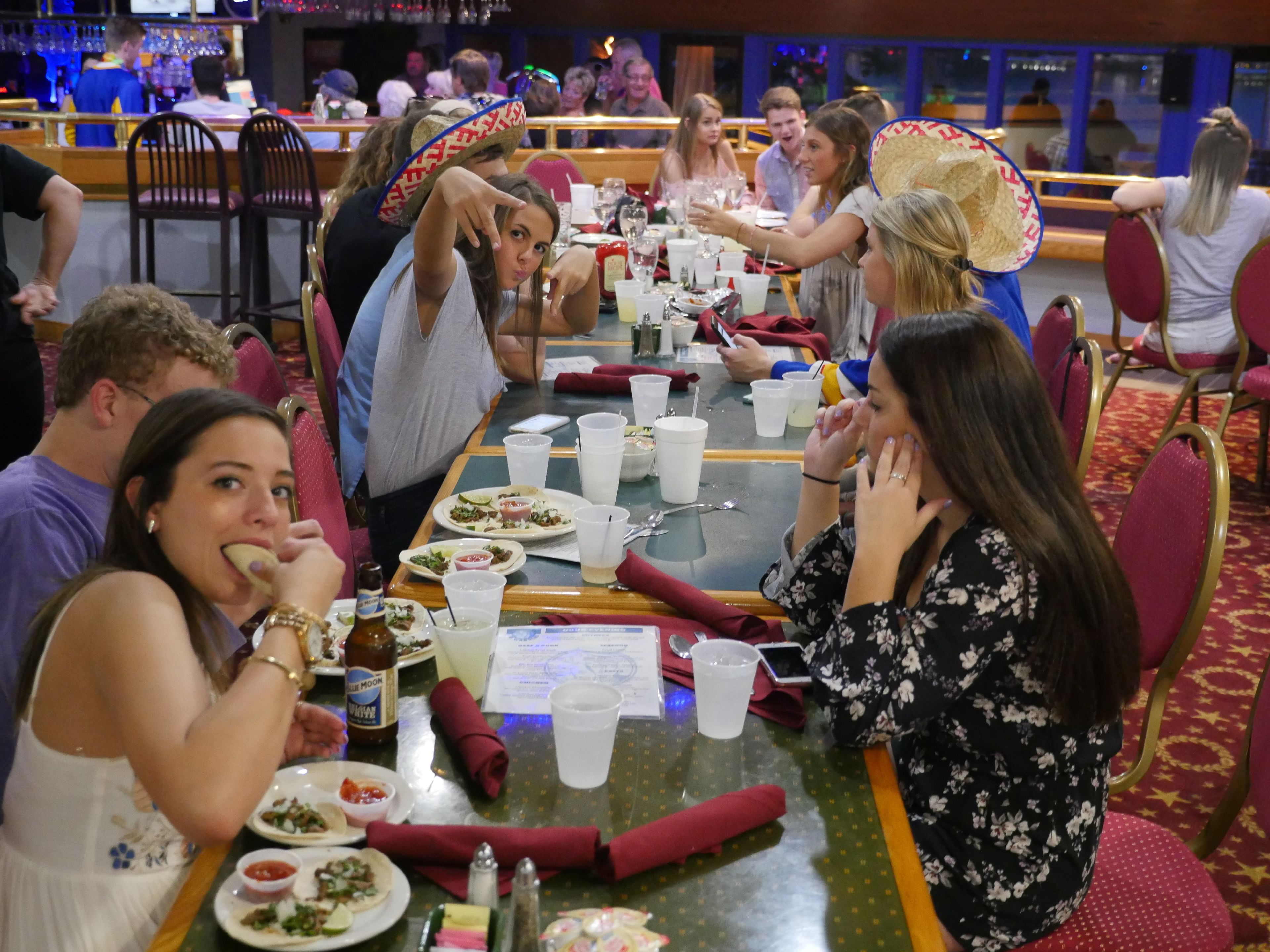 Un grupo de personas cenando en un restaurante con una larga mesa, sillas y diversos platos y bebidas.