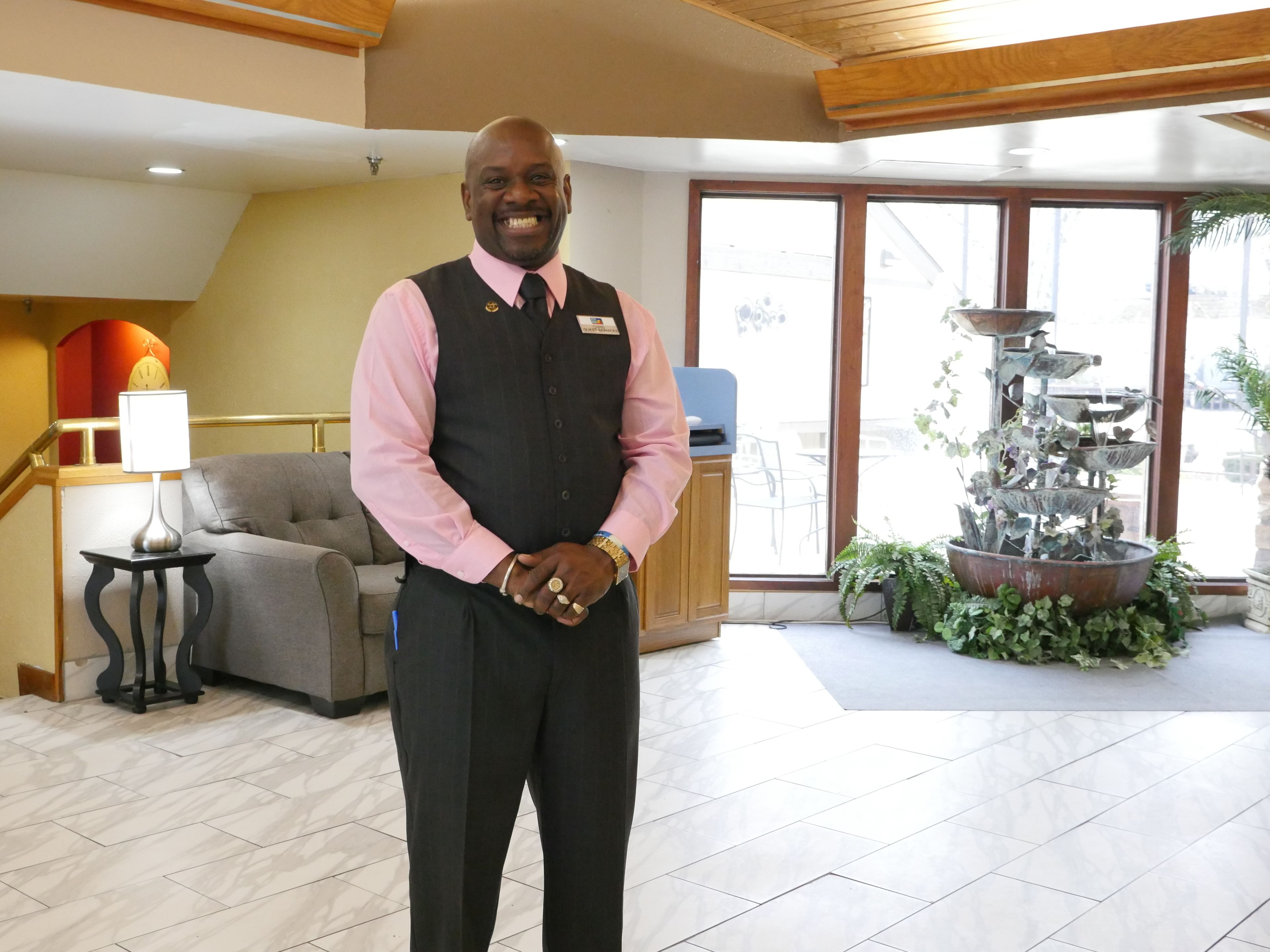 A man in a hotel lobby with a reception desk, a grey sofa, a clock, and a water feature.