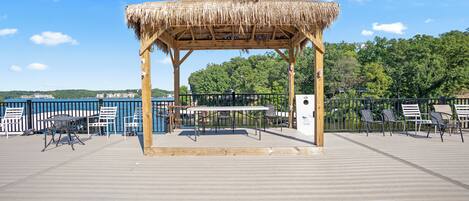A wooden gazebo with a thatched roof on a wooden deck, surrounded by chairs and tables, overlooking a body of water with a view of a forested hill in the background.