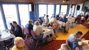 A dining hall with round tables and chairs, large windows, and a patterned carpet.