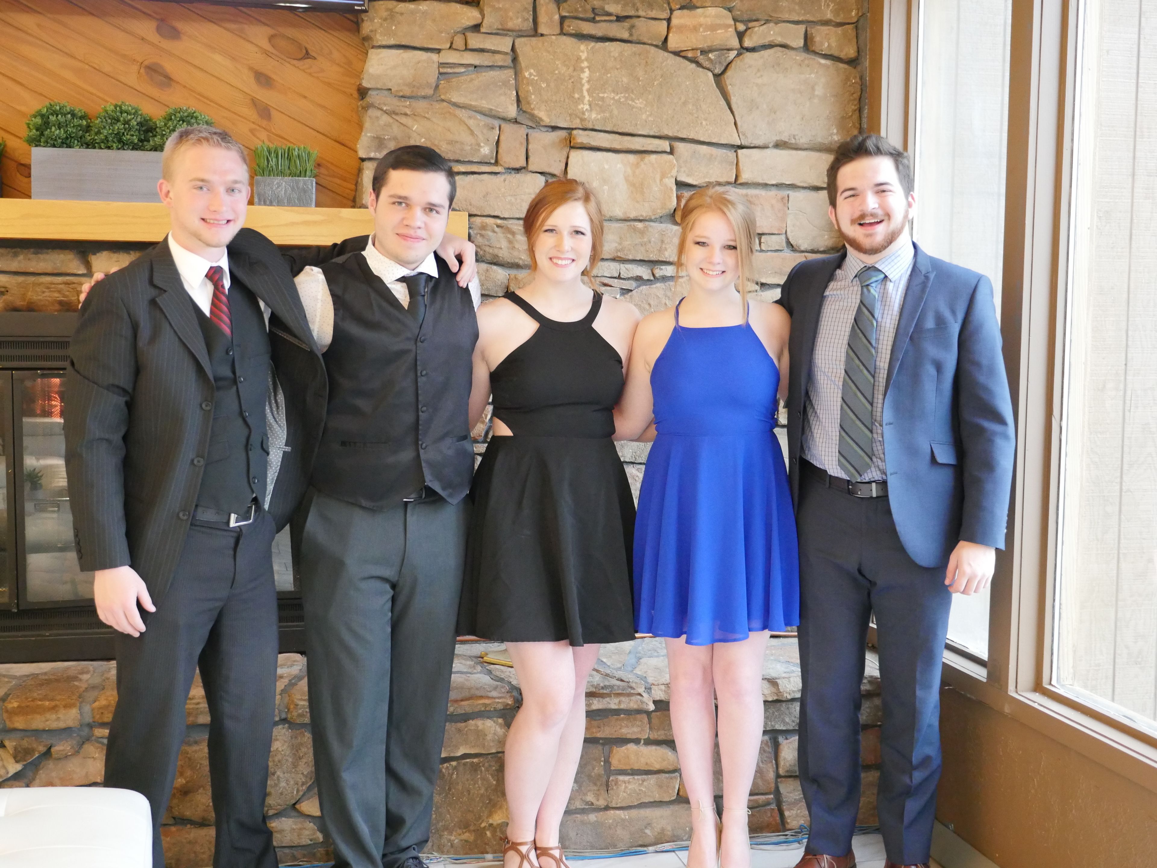 Un grupo de personas vestidas formalmente de pie en un interior con pared de piedra y paneles de madera.