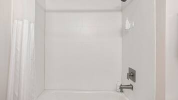 A modern bathroom with white tiles, a shower curtain, a handheld showerhead, and a bottle of shampoo.