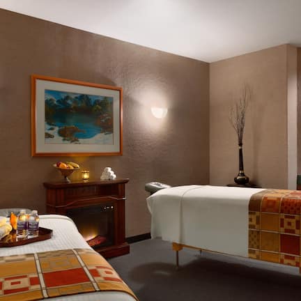 A hotel room with two beds, a fireplace, a painting, and a small table with towels and toiletries.