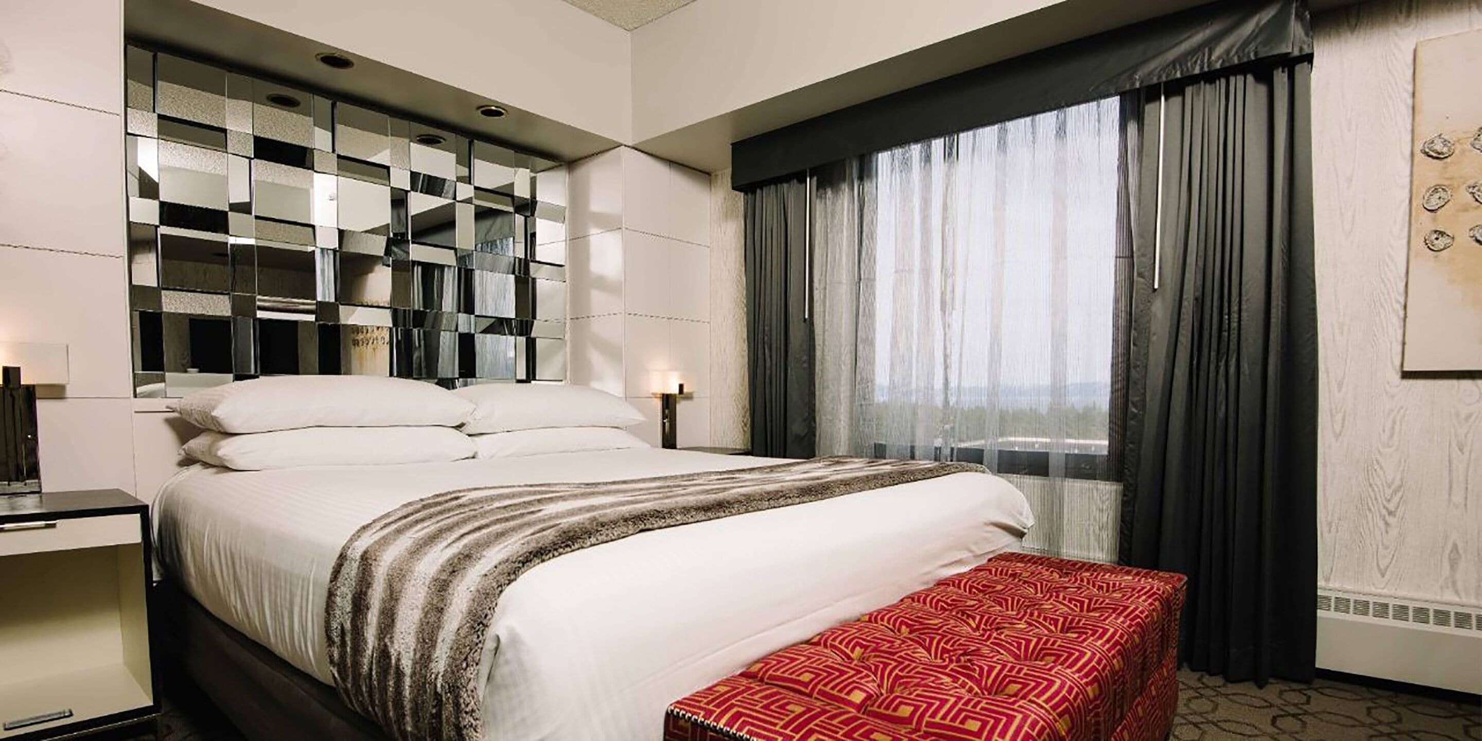 A hotel room with a large bed, a bedside table, a bench, a mirror, and a window with curtains.
