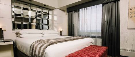 A hotel room with a large bed, a bedside table, a bench, a mirror, and a window with curtains.
