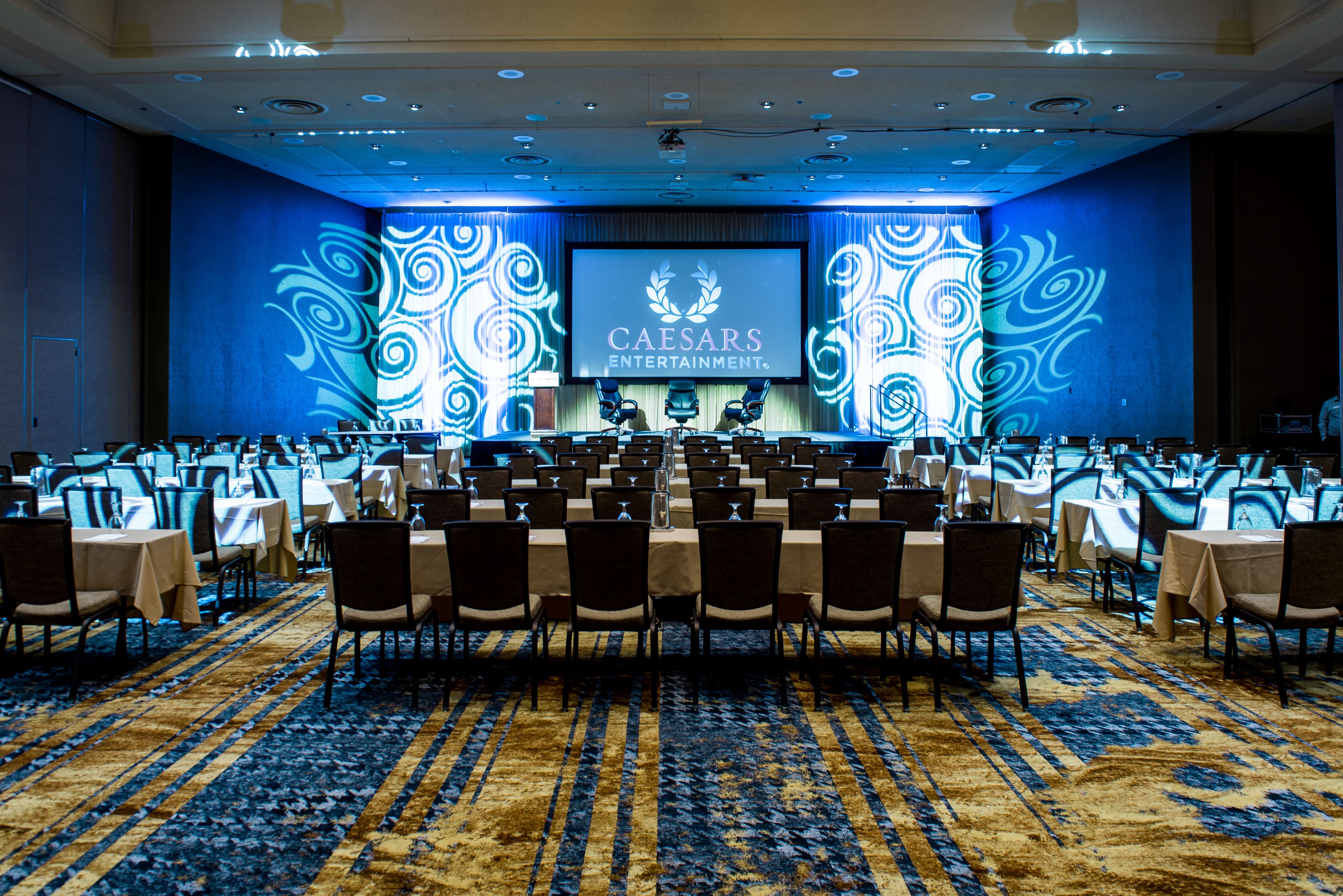 A conference room with a large screen displaying "Caesars Entertainment" logo, rows of tables with chairs, and a patterned carpet.