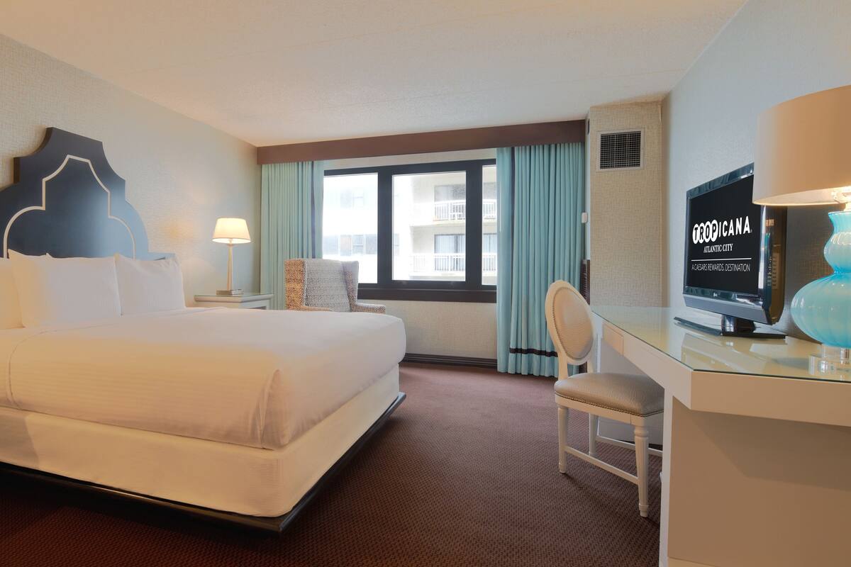Ett hotellrum med en stor säng, ett skrivbord med en tv, en stol och ett fönster med gardiner.