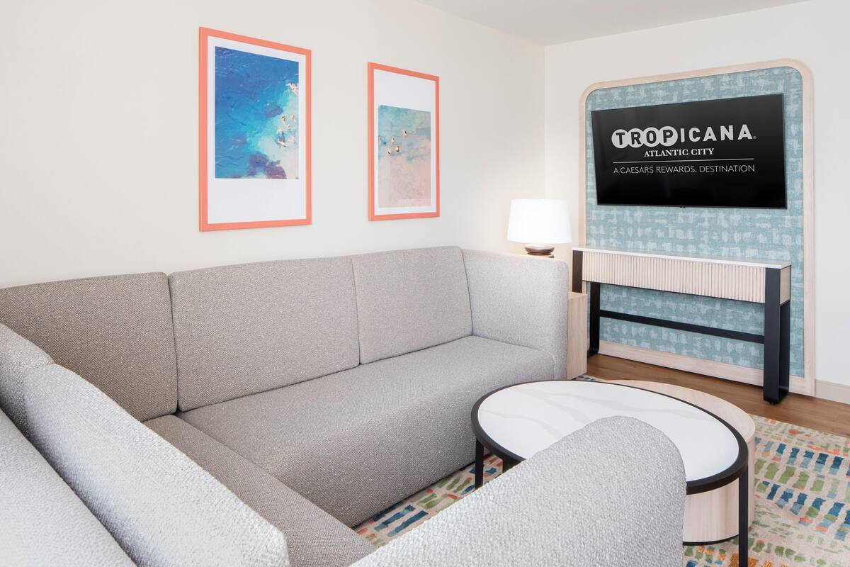 Ett modernt vardagsrum med en grå soffa, ett runt soffbord, inramade konstverk på väggen och en tv-skärm som visar ”TROPICANA ATLANTIC CITY”.
