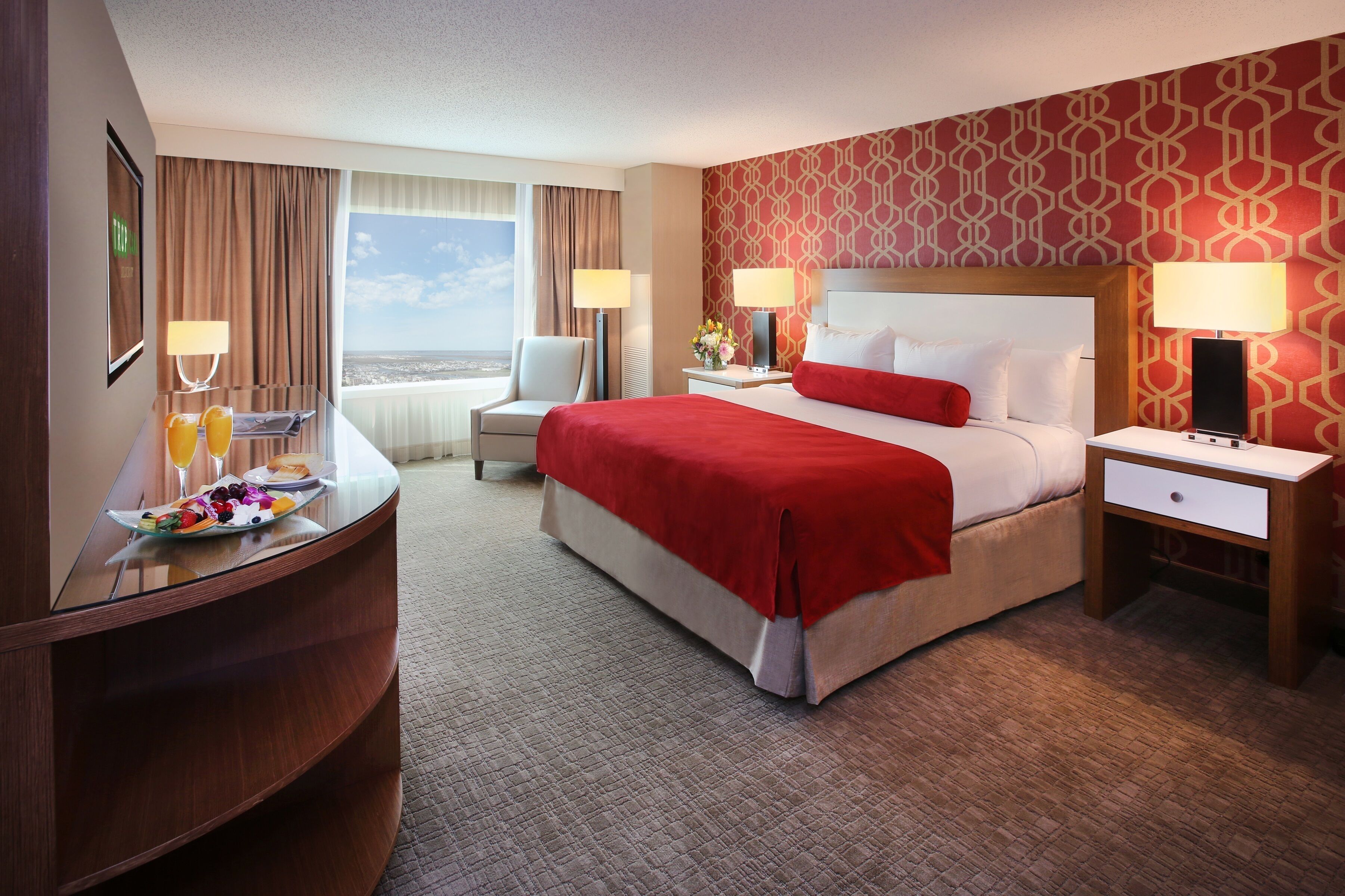 A hotel room with a bed, two bedside tables, a desk with a chair, a large window, and a view of the city.