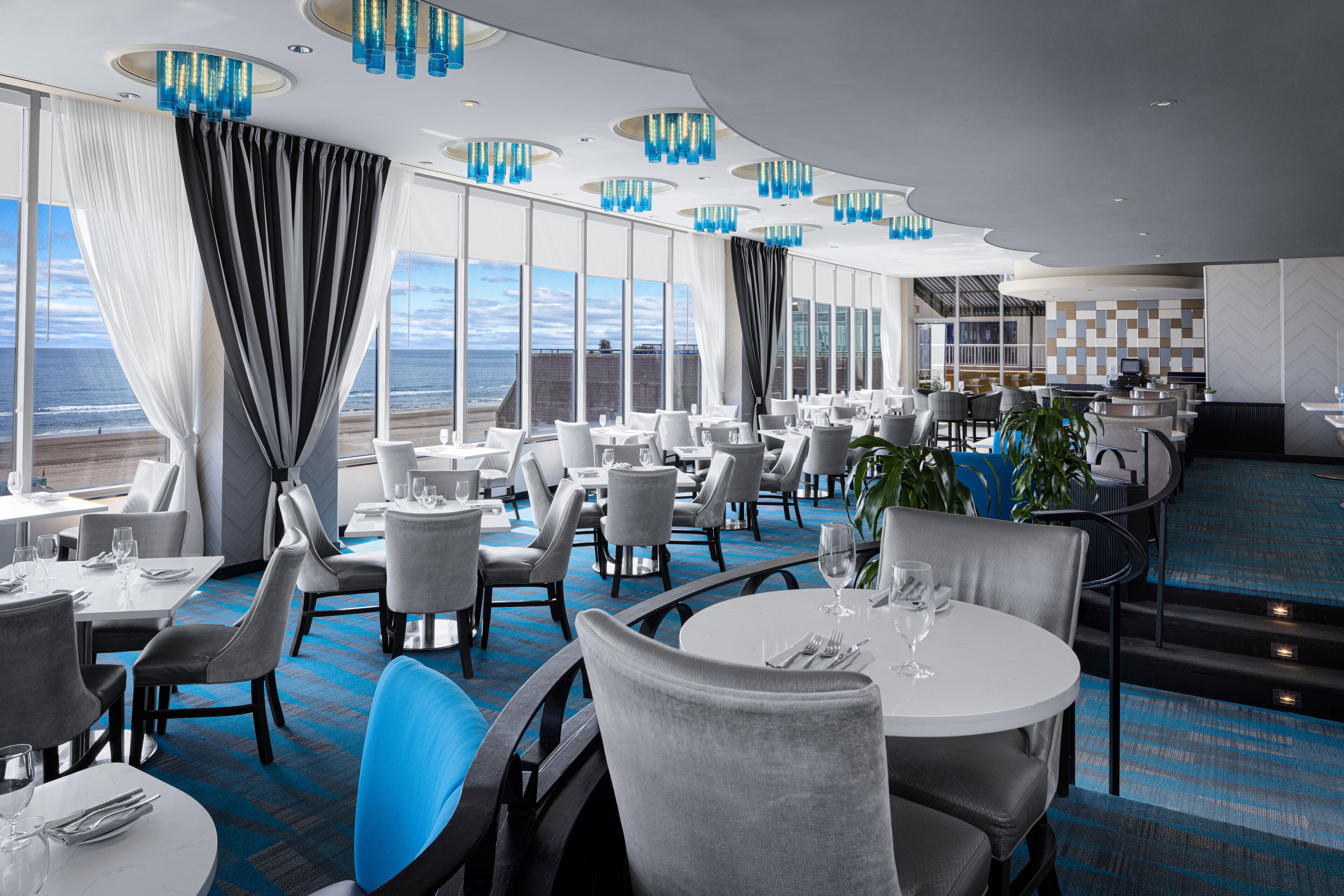 Un restaurante moderno con mesas y sillas redondas, ventanas amplias con cortinas y vistas al océano.