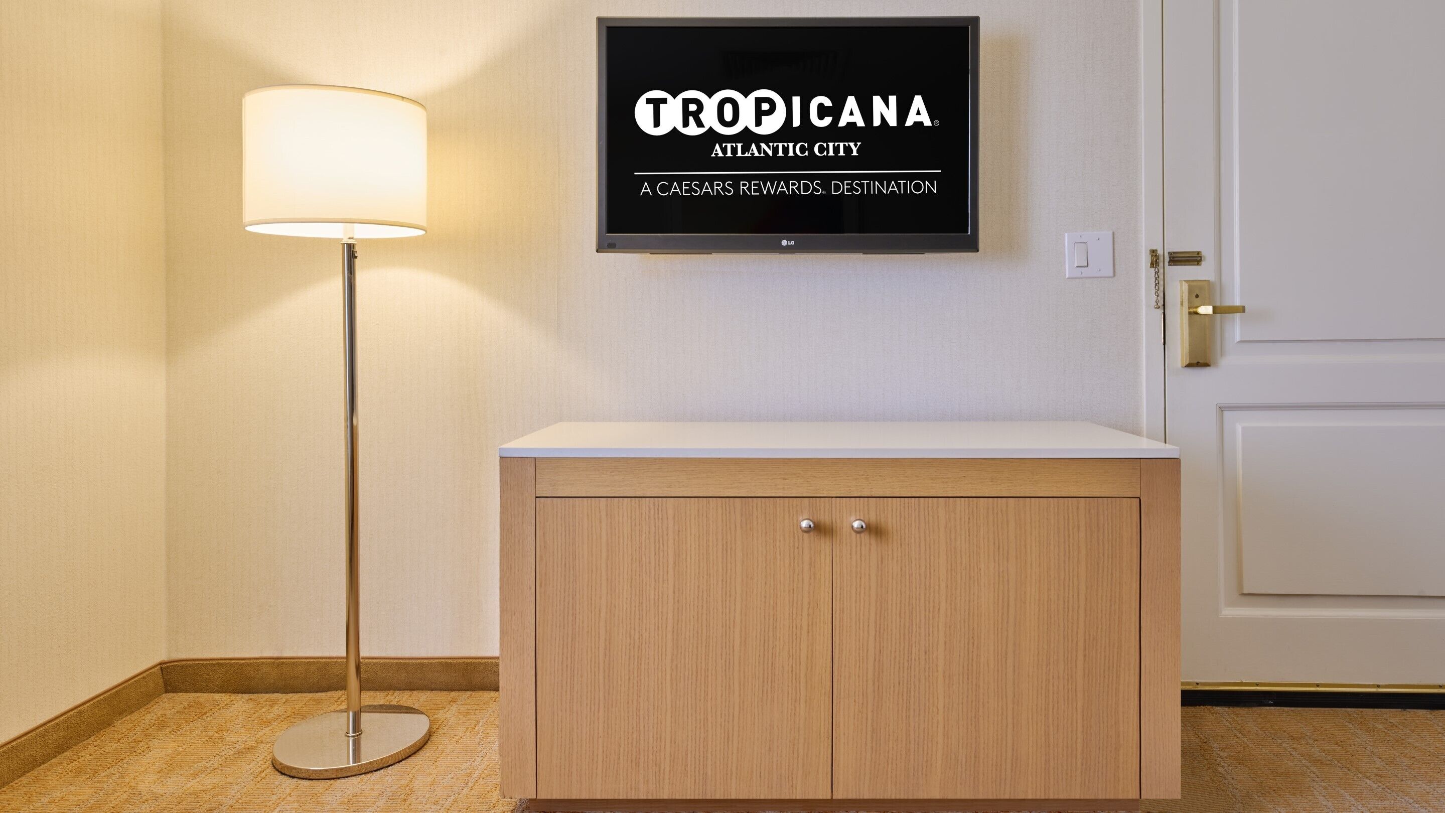 A hotel room with a wooden cabinet, a floor lamp, and a television displaying 'TROPICA ATLANTIC CITY'.