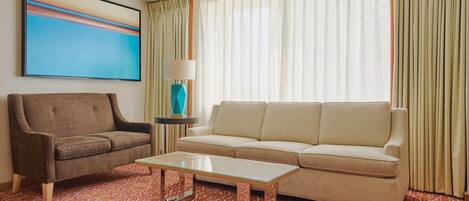 Una sala moderna con un sillón marrón, sofá color beige y una mesa de centro de vidrio. Hay un cuadro en la pared y una lámpara sobre una mesita.