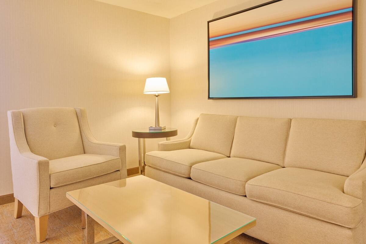 Ett hotellrum med en beige soffa, en tillhörande fåtölj, ett soffbord i glas och en golvlampa.