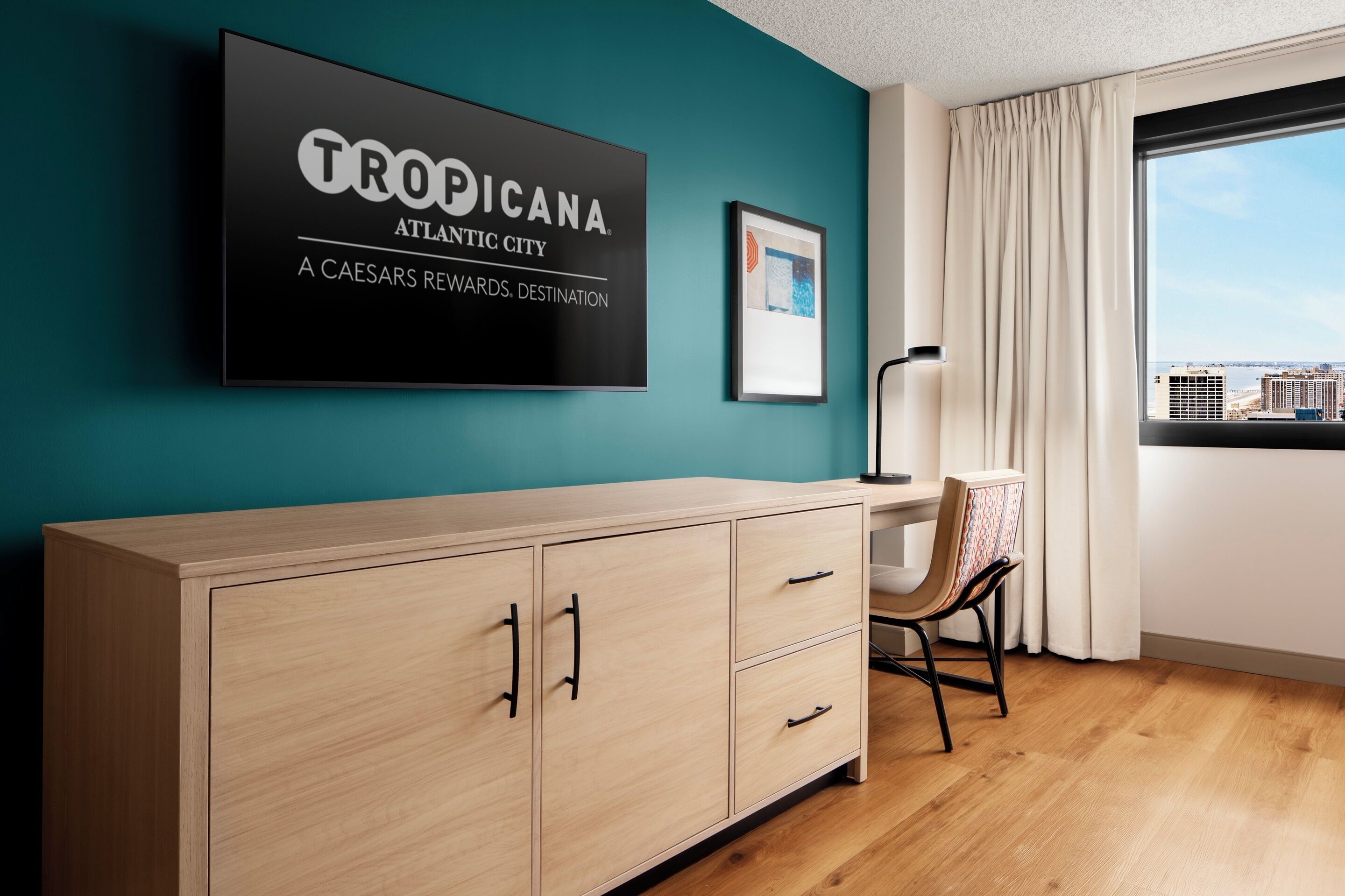 Una habitación con una pared verde azulado, un mueble alto con cajones, una silla, un cuadro enmarco y una ventana con vistas a la ciudad.
