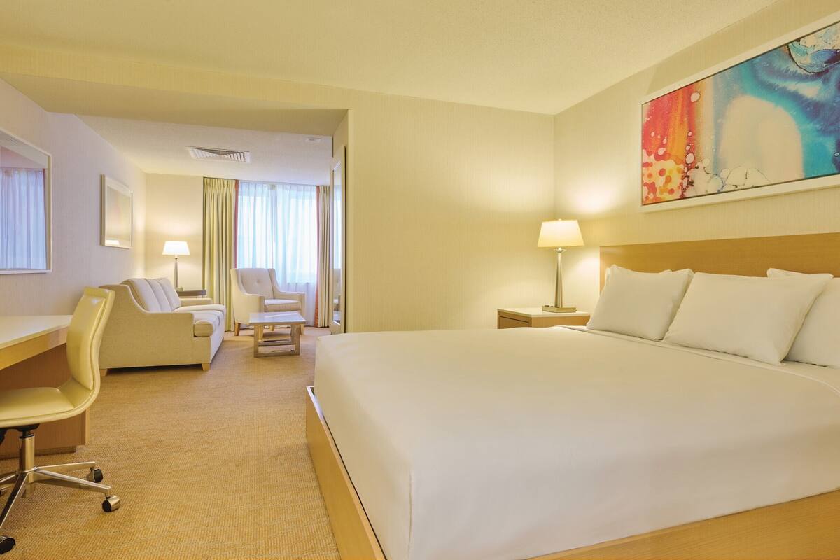Ett hotellrum med en stor säng, ett skrivbord med en stol, en separat fåtölj och en tavla på väggen.