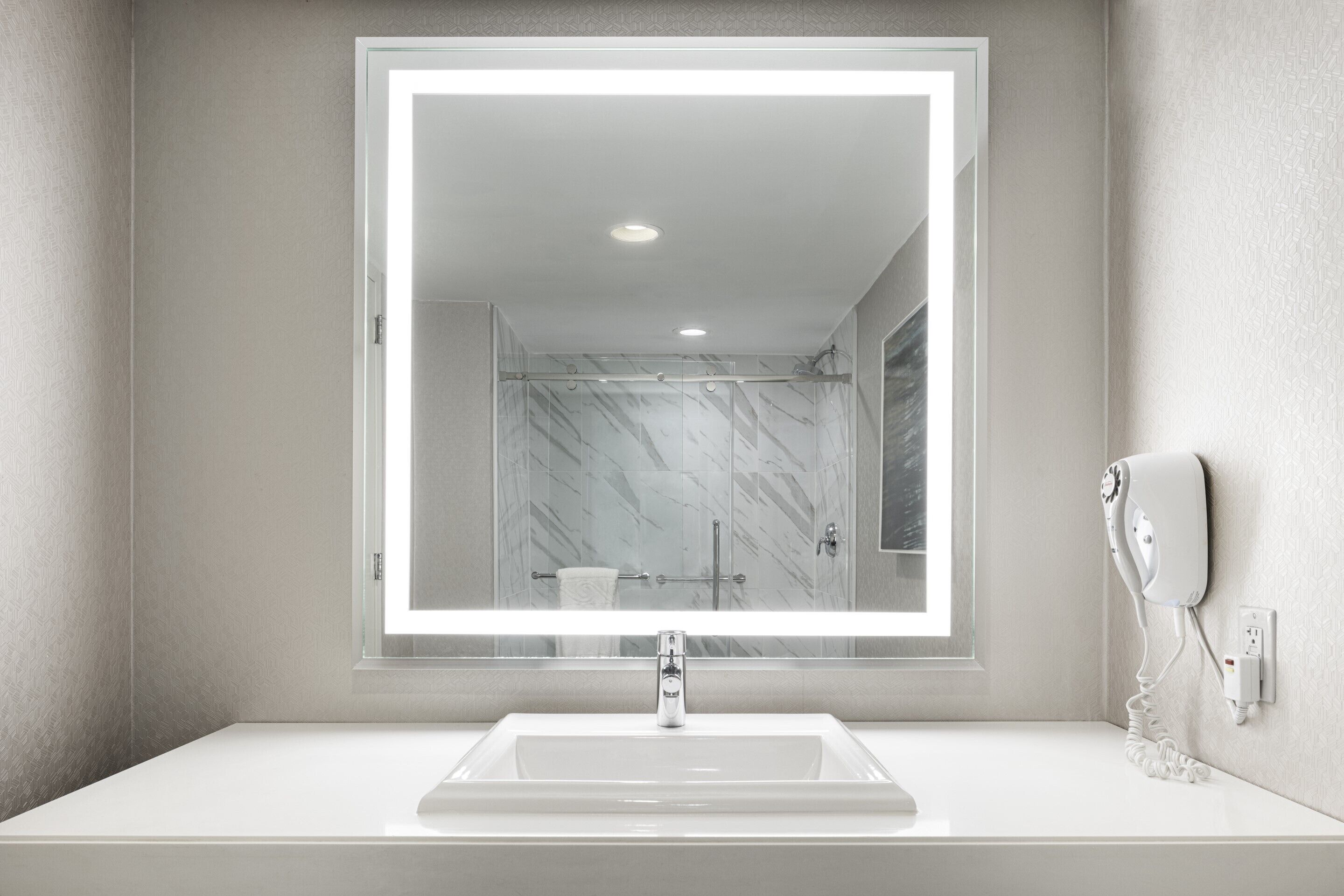 A bathroom with a large mirror, a sink, a handheld hair dryer, and a shower.