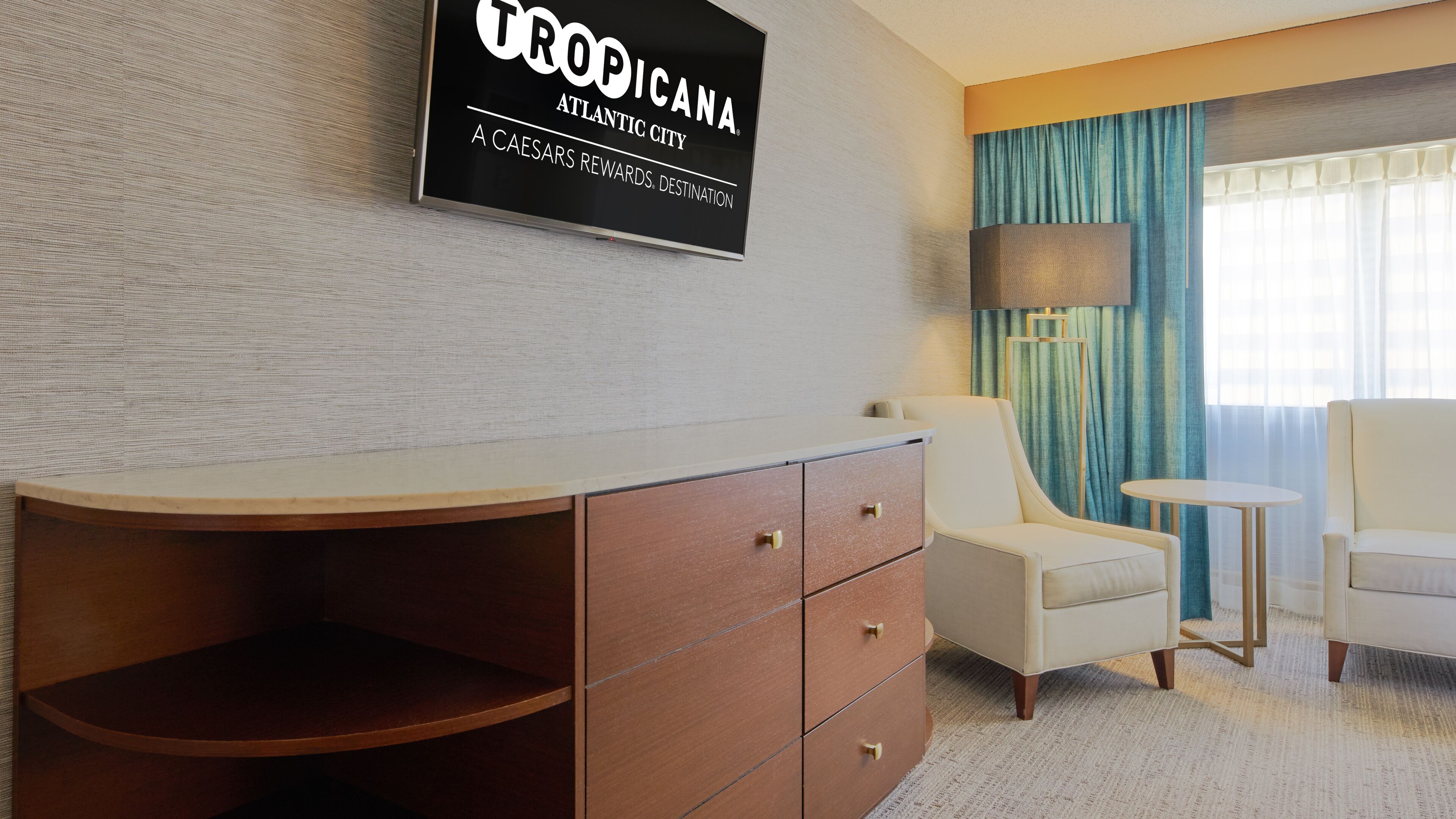 A hotel room with a wooden cabinet, a white armchair, a small round table, and a large window with blue curtains.