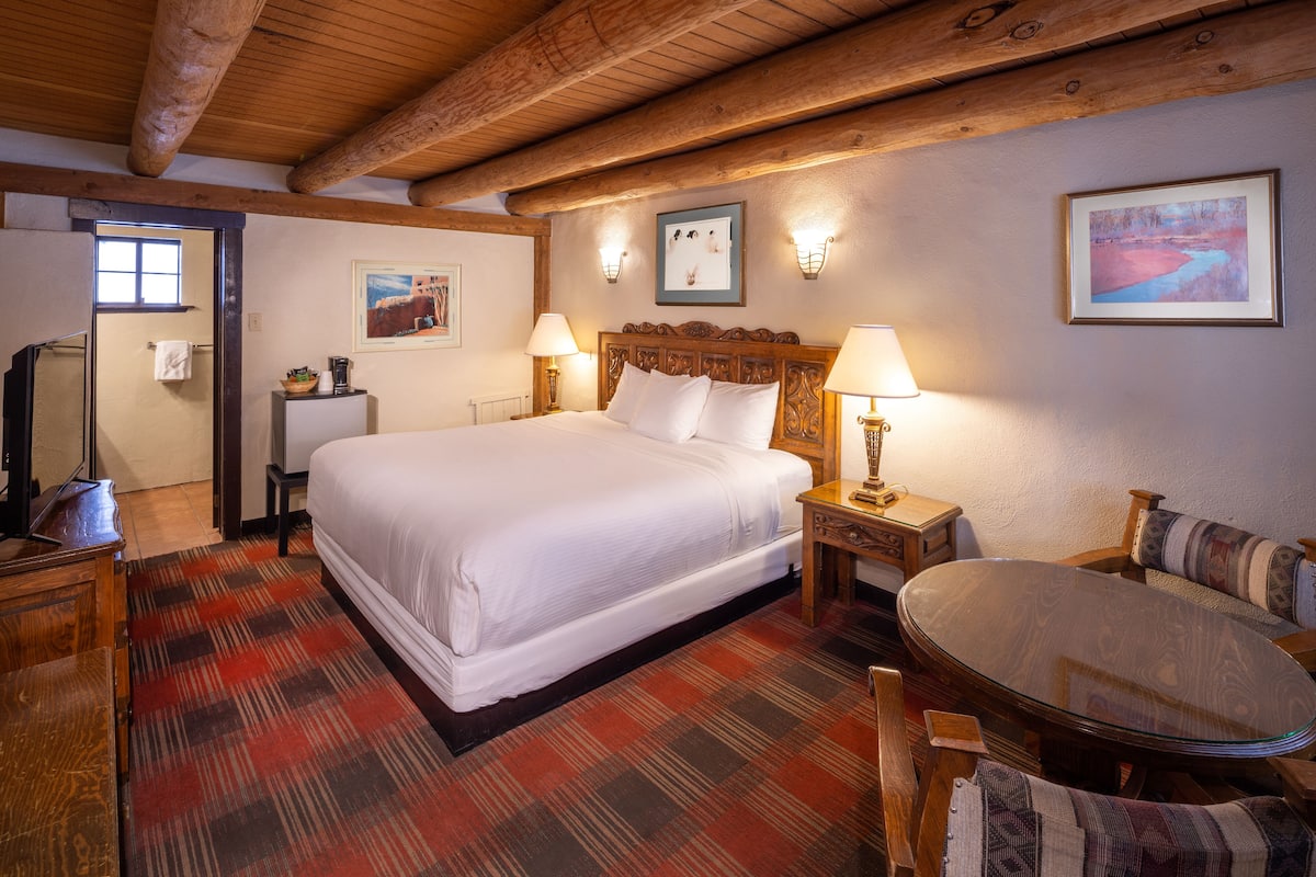 A bedroom with a large bed, wooden headboard, two bedside lamps, a small table with a lamp, a chair, and a painting on the wall.