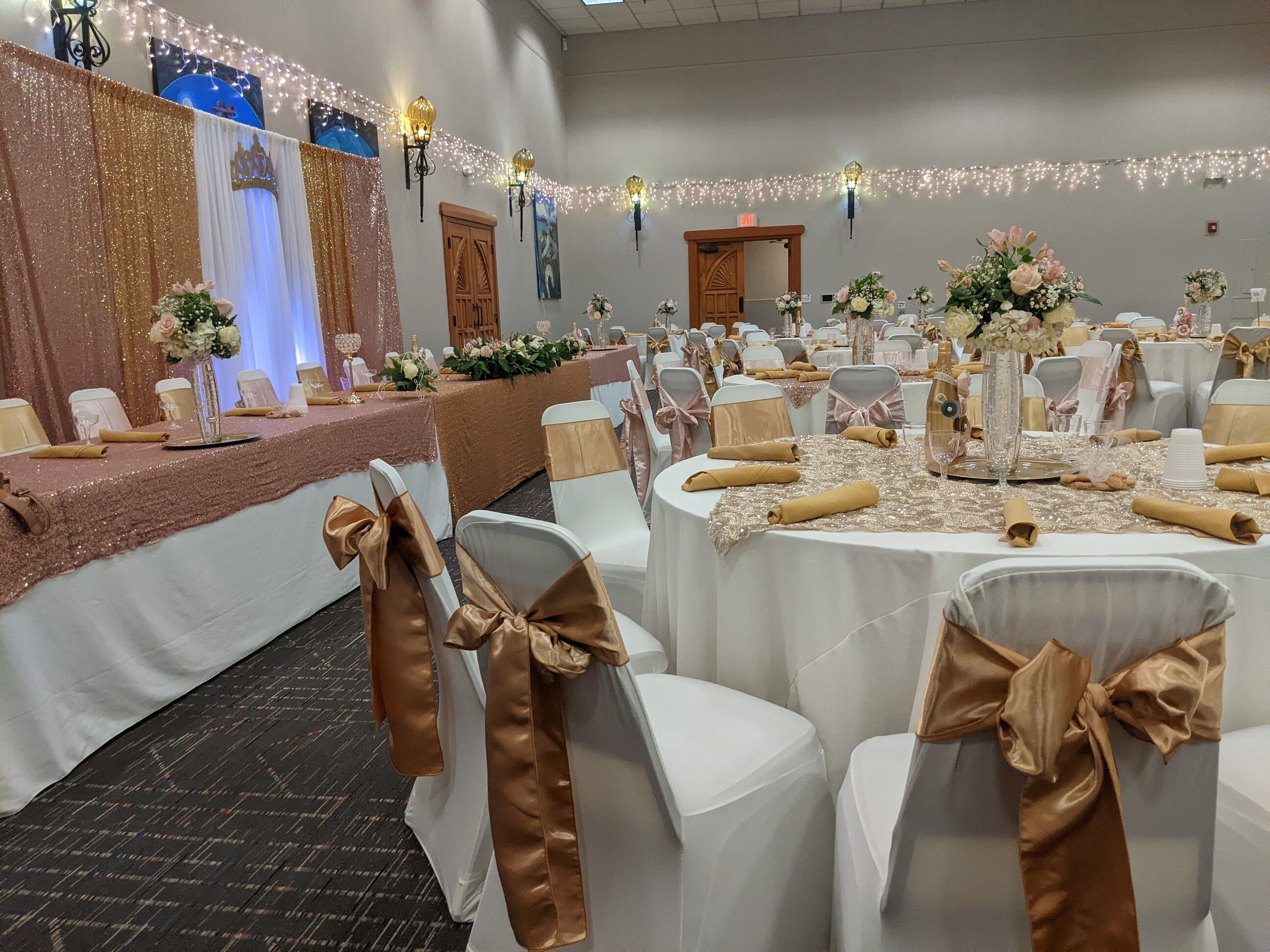 A banquet hall with round tables covered in white tablecloths, gold sashes on chairs, and floral centerpieces.