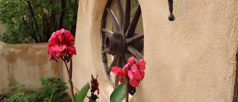 A rustic building with a wheel-like structure, surrounded by greenery and pink flowers.