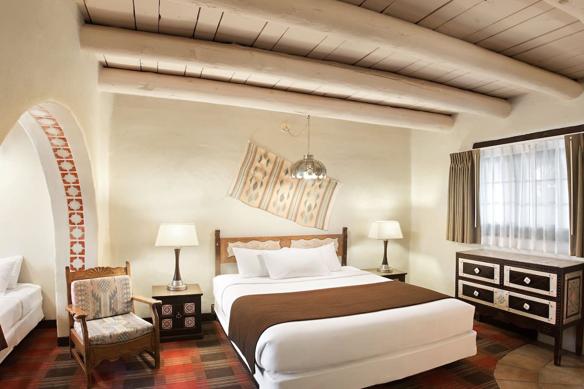 A hotel room with a large bed, two bedside tables with lamps, a wooden chair, a patterned wall hanging, and a window with curtains.